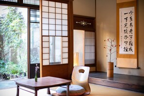 A traditional Japanese room with tatami flooring, a low table, a cushion, a wooden chair, and a sliding door with a view of greenery outside. - Higurashi-Sou (Kyoto)