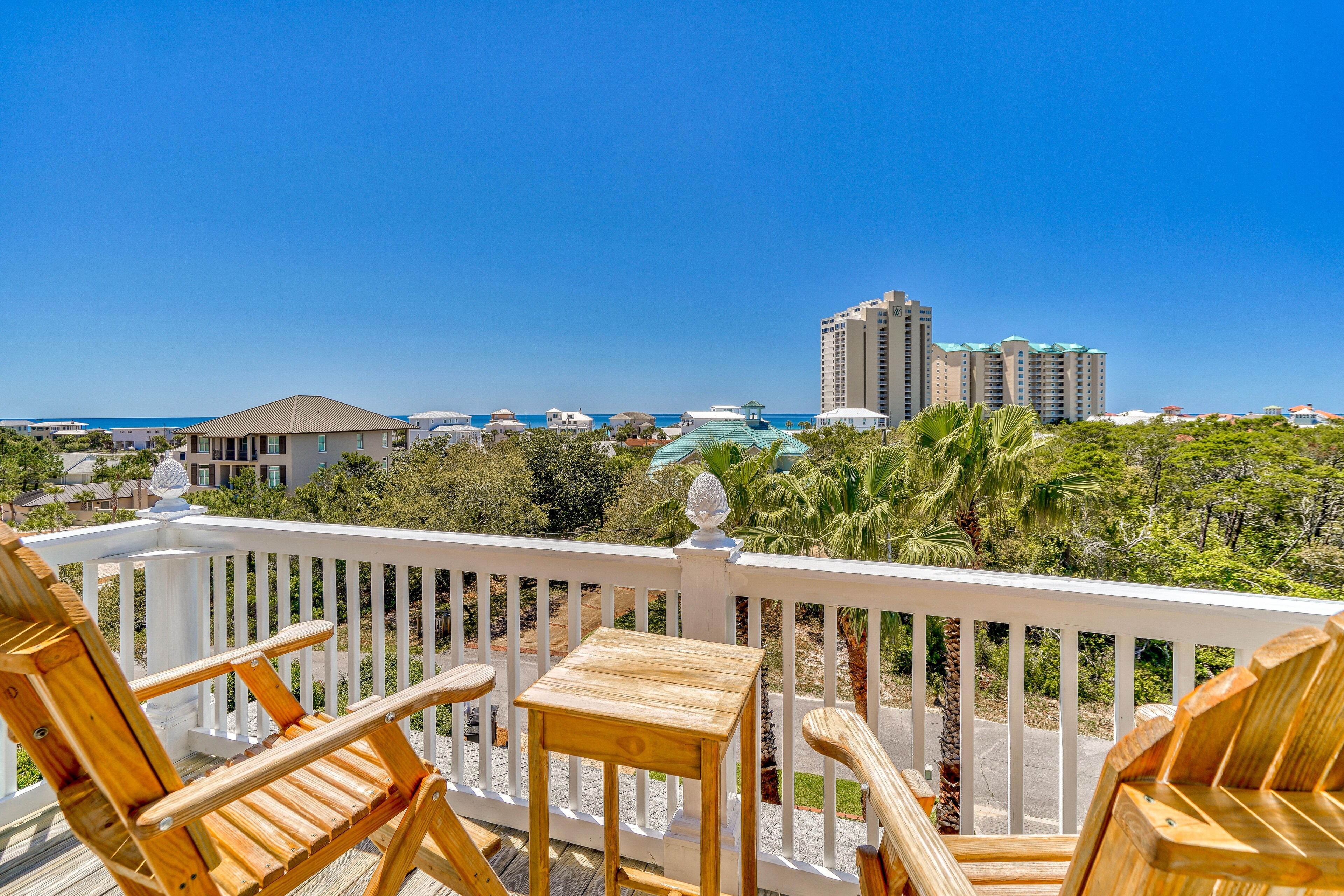A wooden deck with chairs and a table, overlooking a beach and buildings.