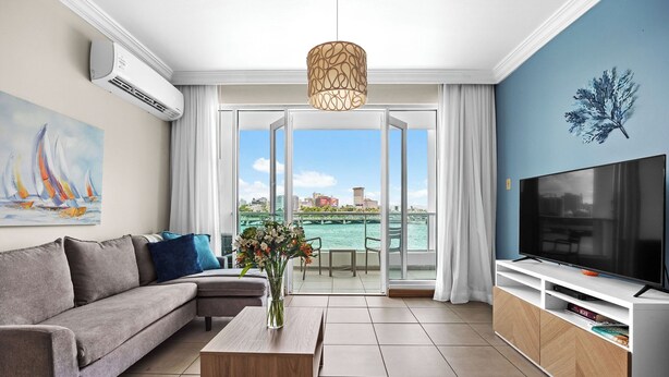 A modern living room with a grey sofa, a wooden coffee table, a flat-screen TV on a white stand, and a view of the water through large windows.