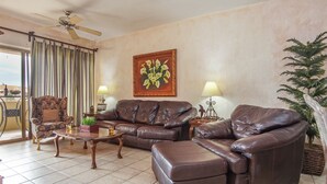 Una sala de estar con un sofá de cuero marrón, una mesa de centro de madera, un sillón estampado y un cuadro de flores amarillas en la pared.
