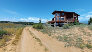 A two-story wooden cabin with a balcony, surrounded by a dirt road and sparse vegetation. - Juniper Ridge 5 BR Family Cabin (Blanding)