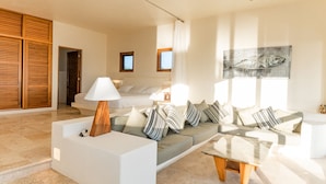 A modern living room with a sofa, a coffee table, and a lamp. There is a bed with a window and a painting on the wall.