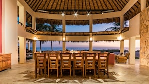 A spacious dining area with a thatched roof, a large table, and chairs, overlooking a scenic ocean view.