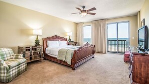 A hotel room with a large bed, a television, a balcony with a view of the ocean, and a ceiling fan.
