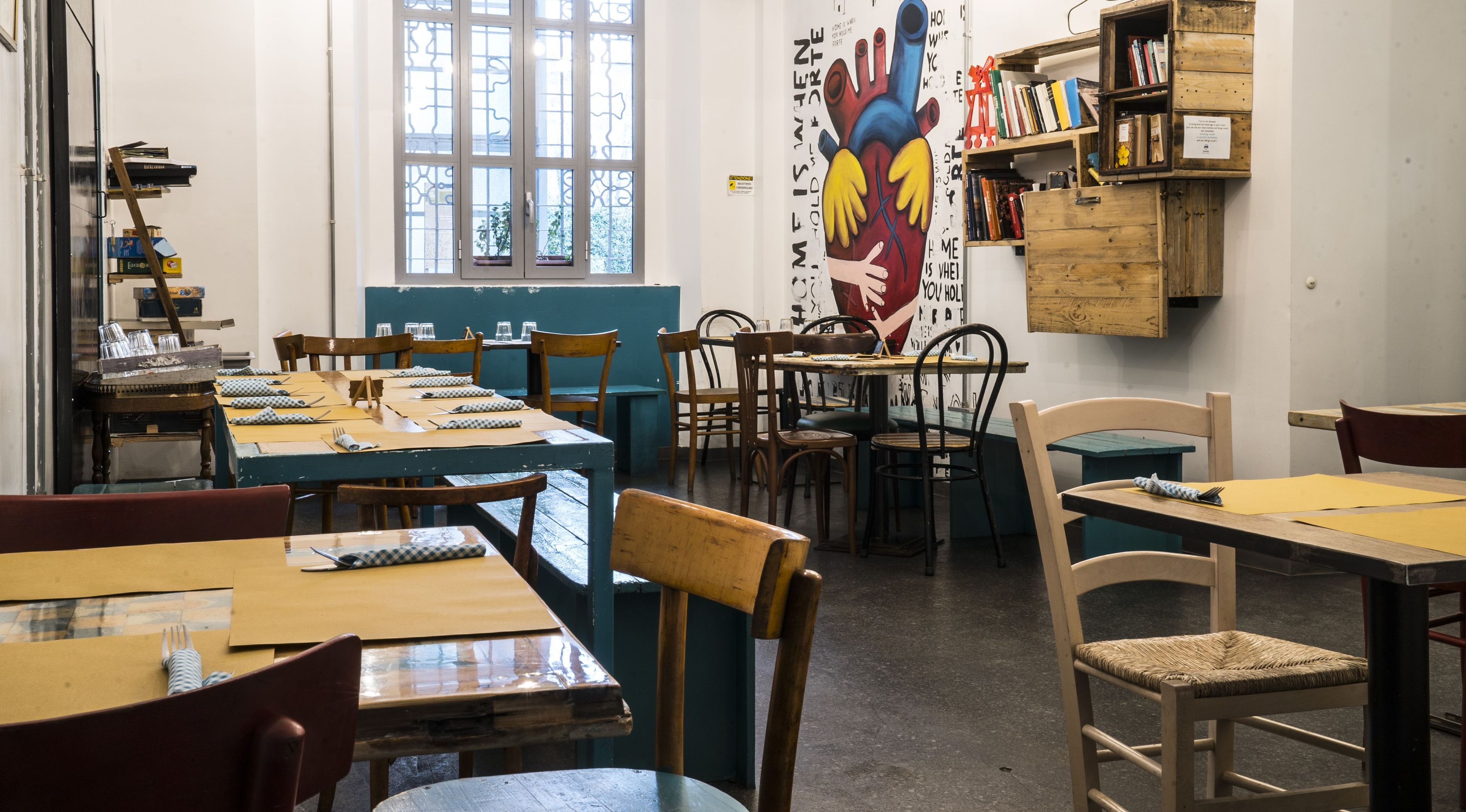 Un comedor con mesas y sillas de madera, un mural en la pared y una estantería llena de libros.
