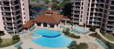 A hotel with a central swimming pool surrounded by sun loungers, a tennis court, and a small building with a red roof.