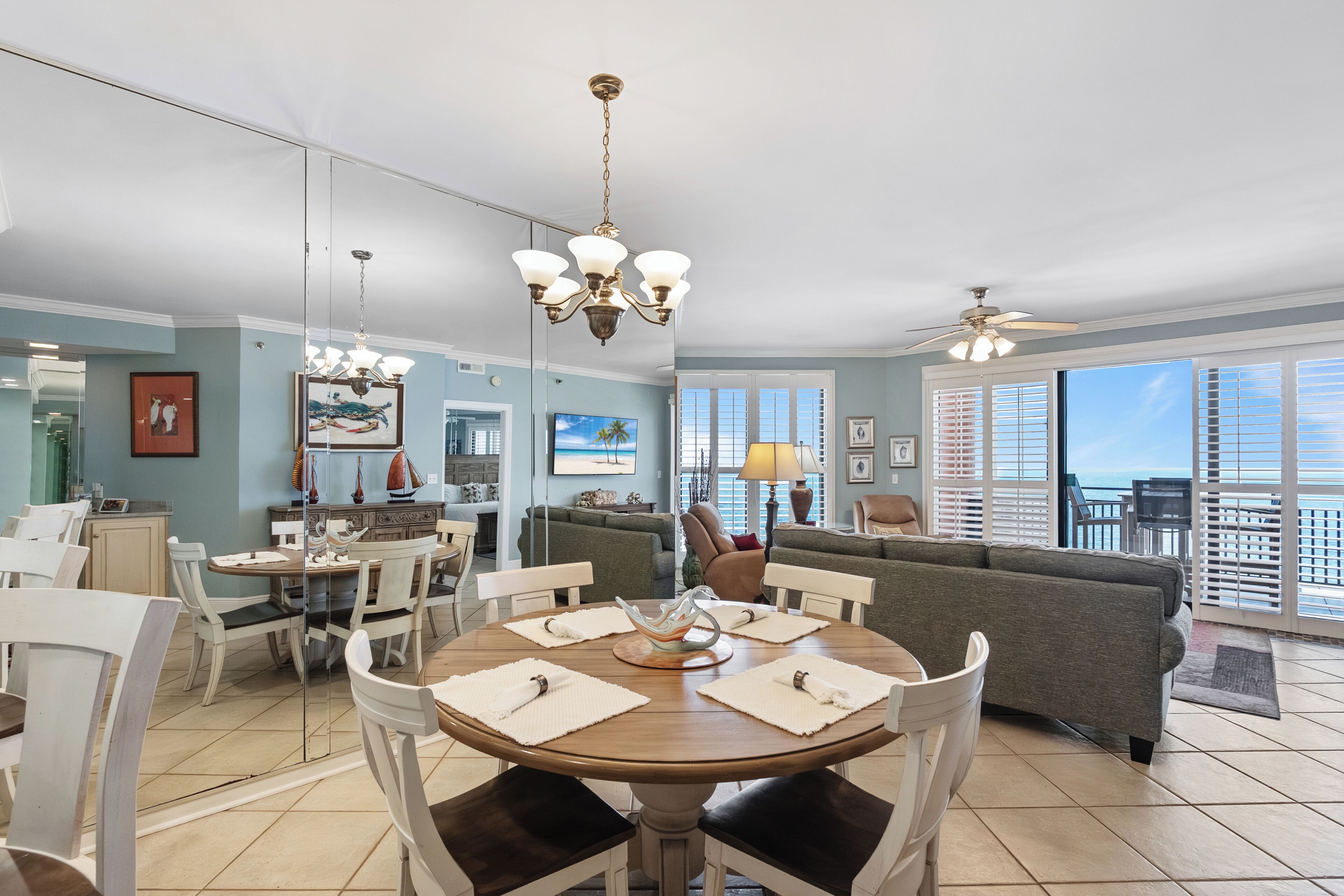 A dining area with a round table, chairs, and a chandelier. There is a sofa and a balcony with a view.