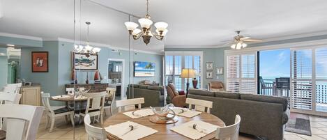 A dining area with a round table, chairs, and a chandelier. There is a sofa and a balcony with a view.