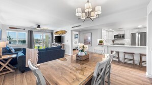 A modern living room with a wooden dining table, blue sofa, and a kitchen area with stainless steel appliances.