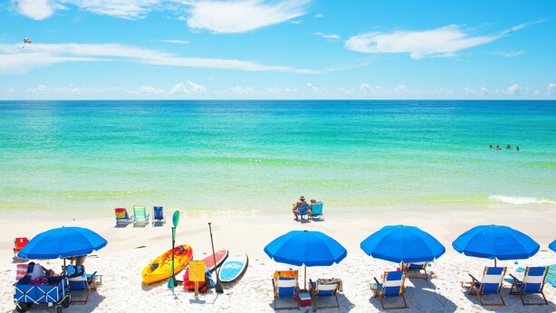 A beach with clear turquoise water, several blue umbrellas, and beach chairs.