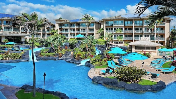 A resort pool area with multiple lounge chairs and umbrellas, surrounded by lush greenery and a multi-story building with balconies. - H206 Ocean View Suite - Modern and Clean - Sleeps 6 in Air Conditioned Comfort! (Kapaa)