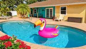 Une piscine avec un flotteur en forme de flamant rose, une maison à la façade turquoise et jaune, et un palmier en arrière-plan.