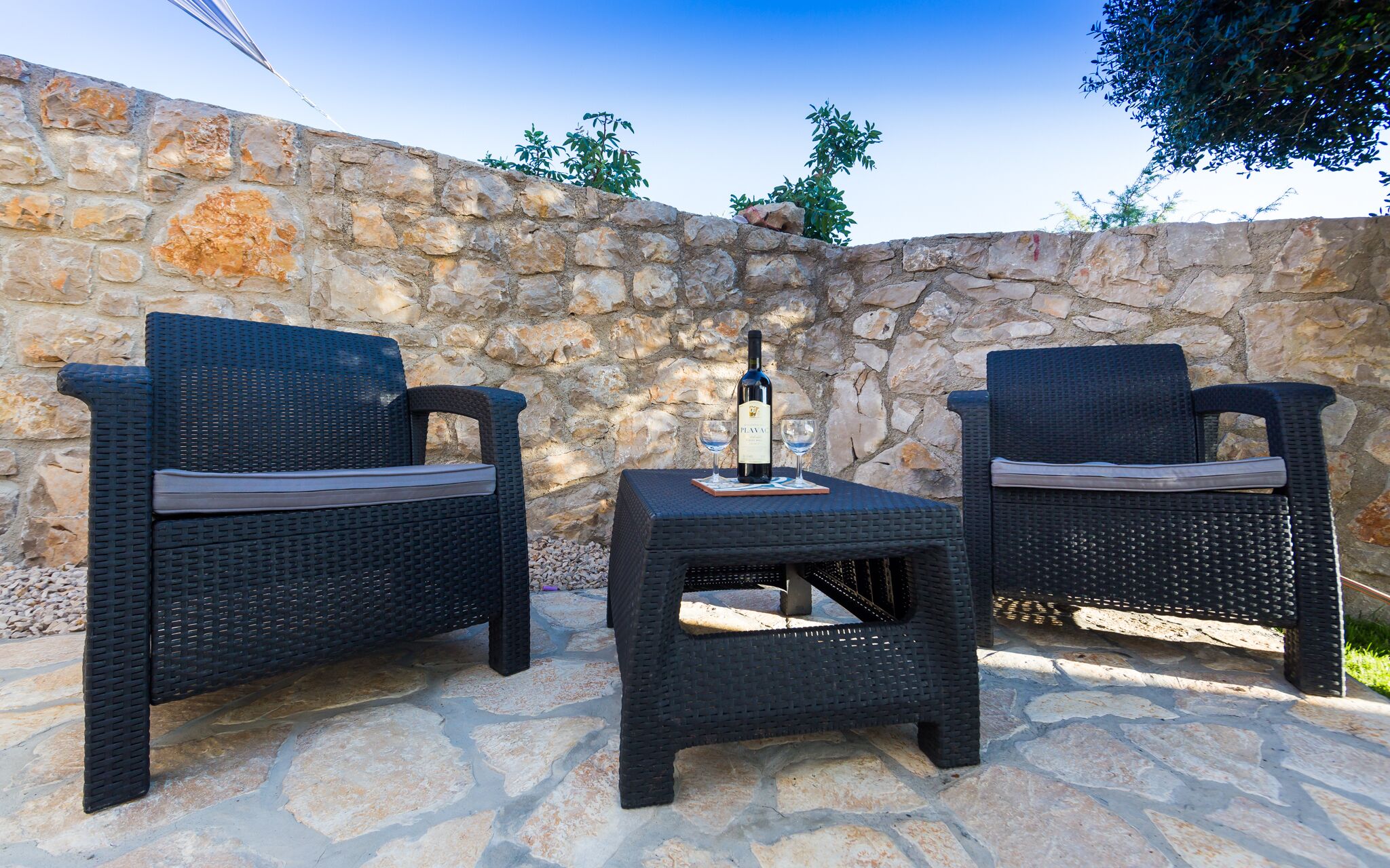 Two wicker chairs with cushions, a small table, and a bottle of wine on a stone patio.