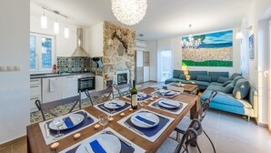 A modern kitchen with a stone wall, a dining area set for six with blue and white tableware, and a living room with a blue sofa and a painting of a landscape. - Hacienda Babina, Island Cres, Lounge, Whirpool, Pool...Seaview (Miholascica)