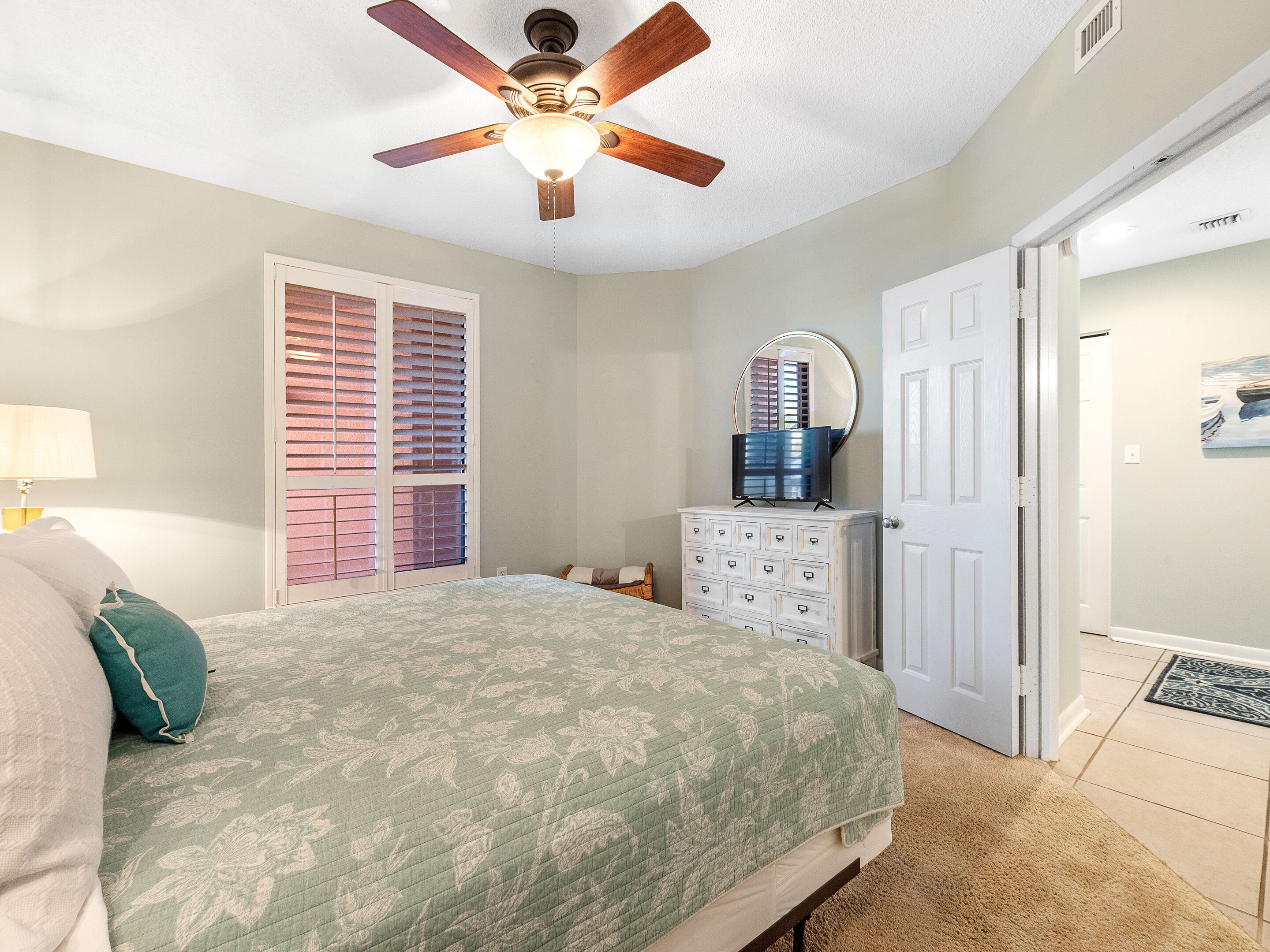 A bedroom with a bed, a dresser, a ceiling fan, a door, and a window with blinds.