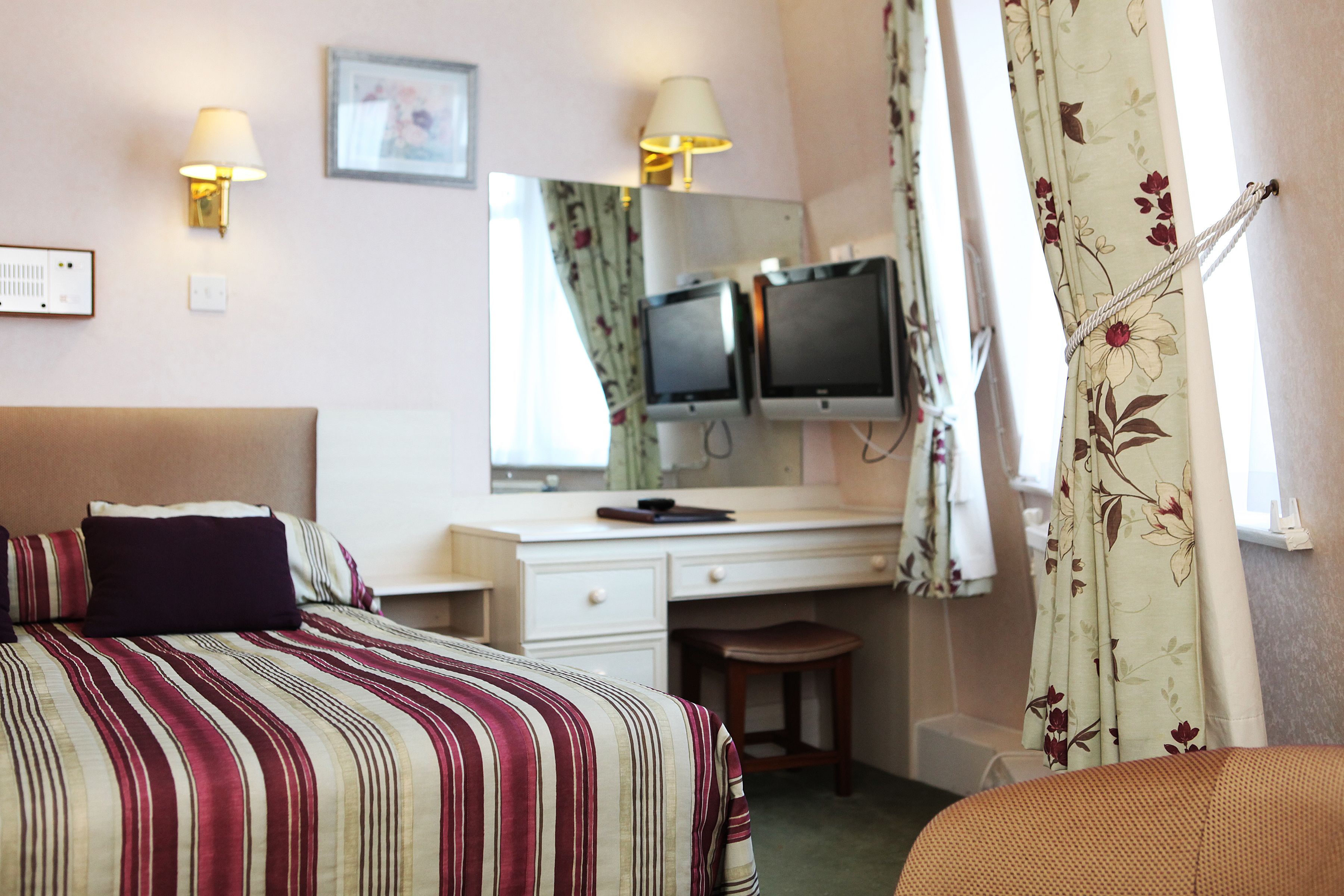 A hotel room with a bed, a desk, two monitors, a chair, and a window with curtains.