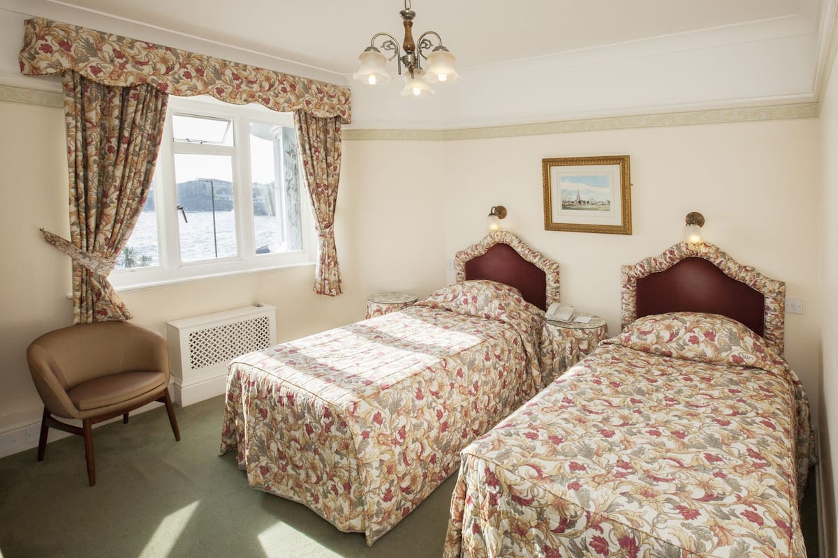 A bedroom with two beds, a chair, a window with curtains, a radiator, and a picture on the wall.