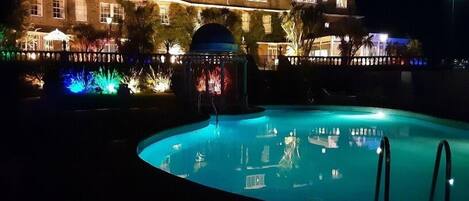 Vue nocturne d’un espace piscine avec un grand bâtiment en arrière-plan.