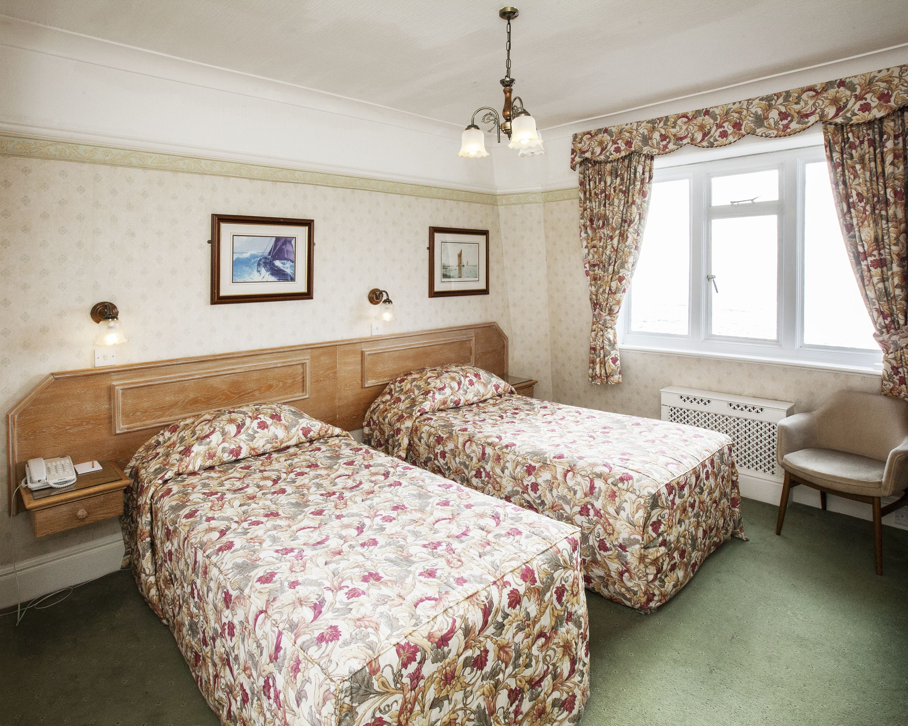 A hotel room with two beds, a chair, a radiator, and two framed pictures on the wall.