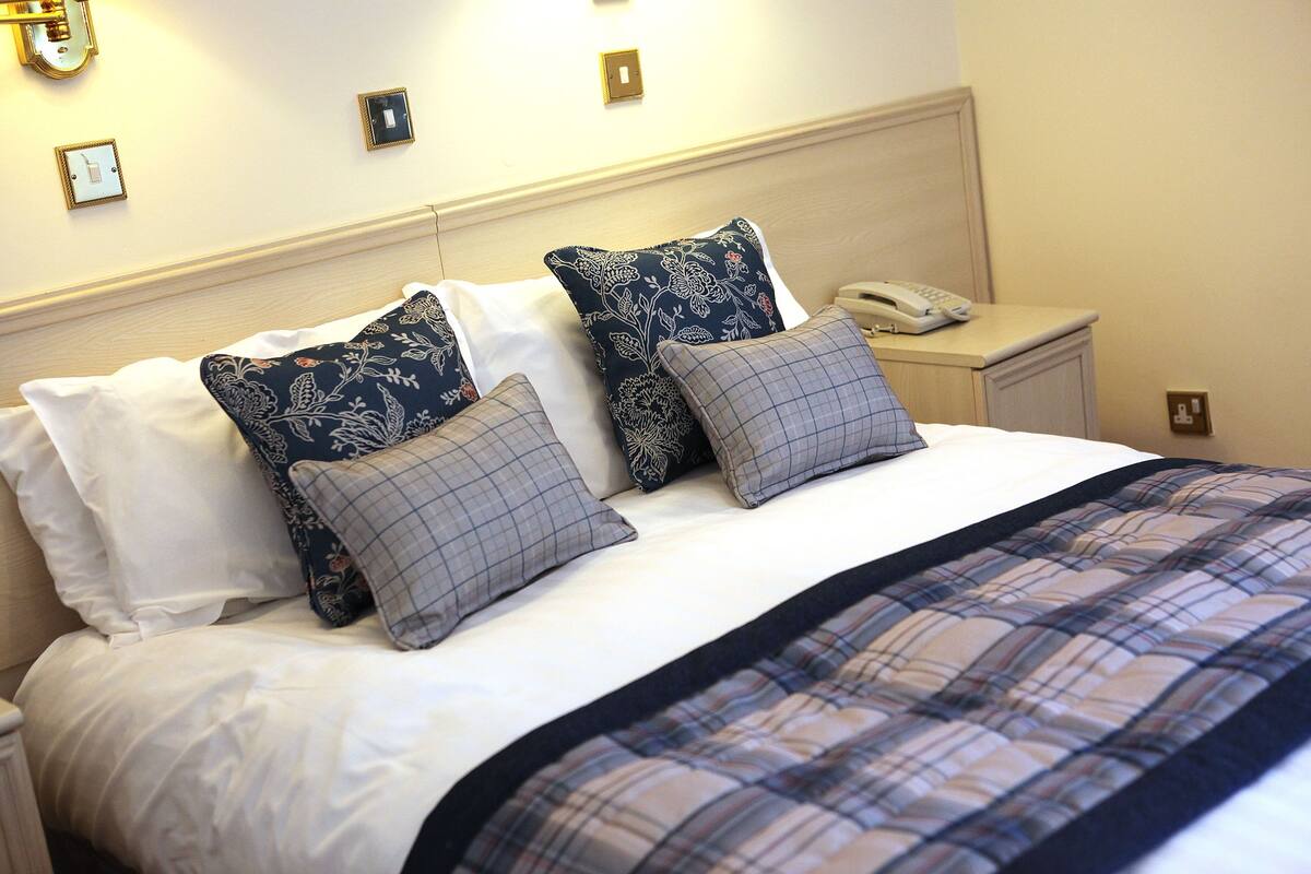 A neatly made bed with a plaid bedspread, two decorative pillows, a bedside table with a phone, and wall-mounted light switches.
