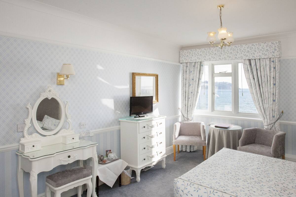 A bedroom with a white vanity, a television, a chair, a bed, and a window with a view.