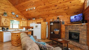 A cozy cabin interior with wooden walls, a kitchen area, a living room with a sofa, a fireplace, and a television.