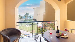 Un balcón con vista a un edificio y palmeras. Hay dos sillas y una mesa con una botella de vino y copas.