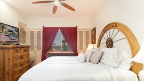 Un dormitorio con una cama, cabecera de madera, ventilador de techo, televisor en una cómoda y una ventana con cortinas rojas.