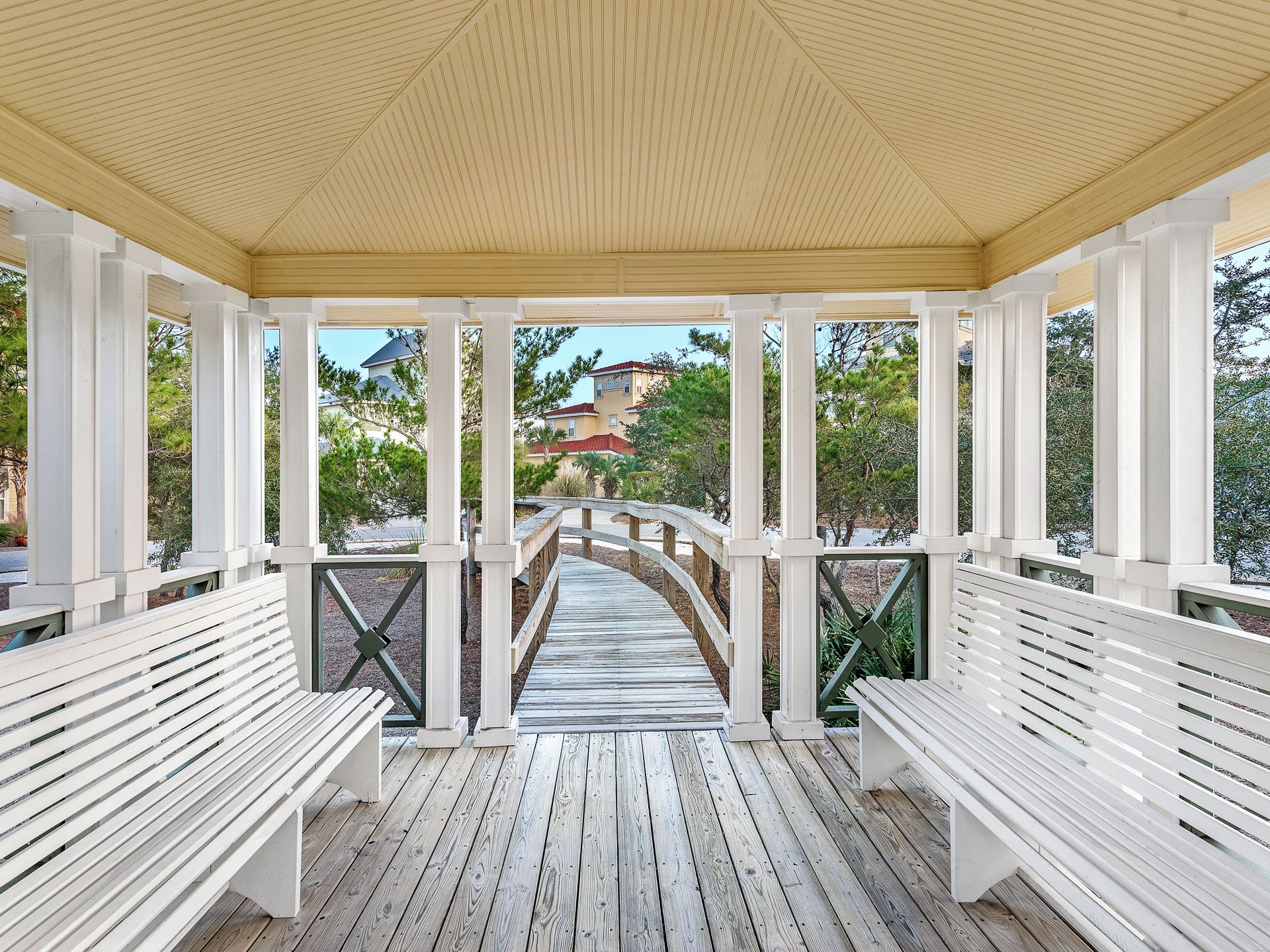 A covered wooden deck with white railings and benches, leading to a bridge over a body of water.