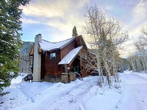 Una cabaña rústica de troncos con techo empinado, rodeada de árboles cubiertos de nieve y un cielo despejado.