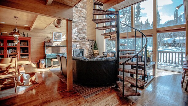A cozy interior with a spiral staircase, wooden floors, a stone fireplace, and large windows offering a view of the outdoors. - Storm Mountain Escape by Cottonwood Lodging (Brighton)