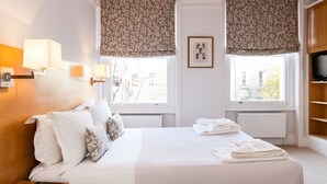 Habitación de hotel con cama, dos ventanas con persianas florales, un televisor y una mesita con un control remoto.