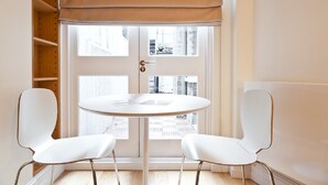 Una habitación con una mesa redonda blanca y dos sillas blancas, una persiana de ventana color beige, y vista a la calle.