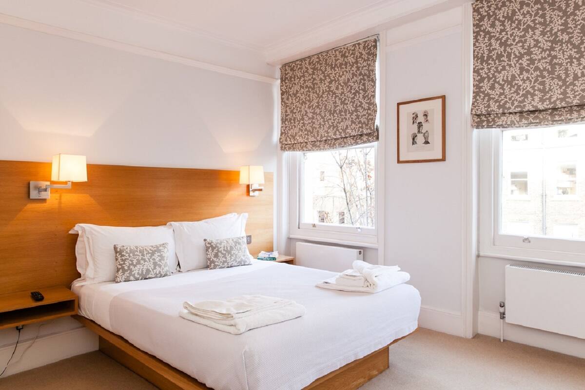 Una cama bien hecha con sábanas blancas y una toalla doblada, un cabecero de madera, dos lámparas de noche, un cuadro en la pared y una ventana con una cortina floral.