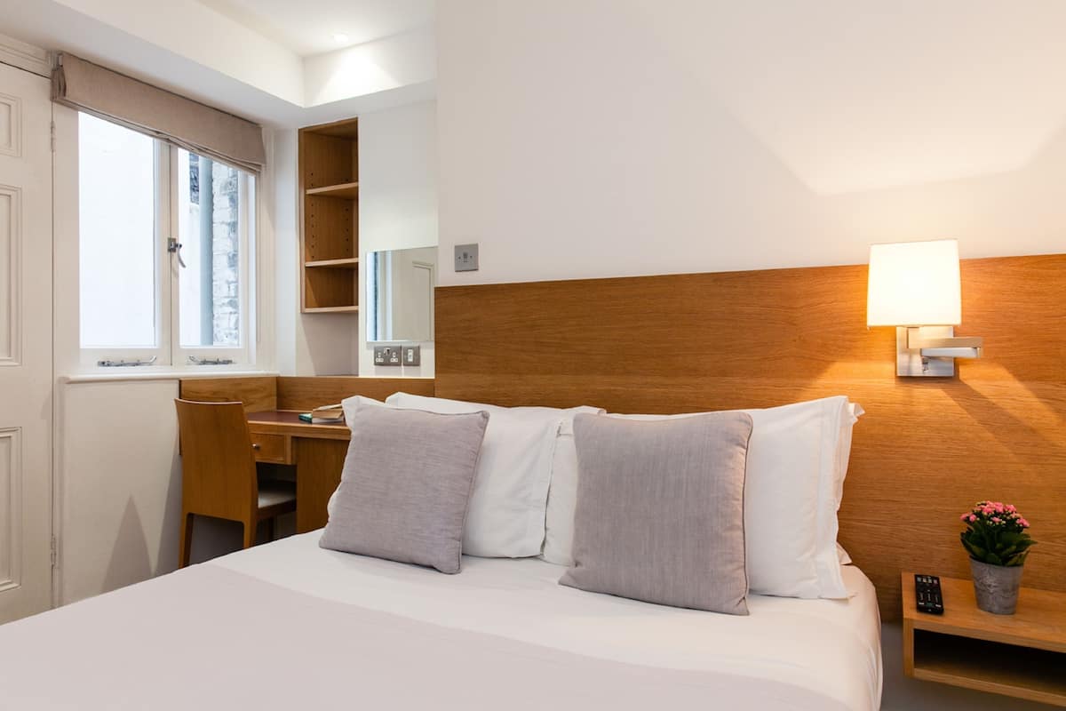 Habitación de hotel con cama, escritorio, silla, mesita de noche, lámpara y ventana.