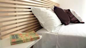 A bed with a wooden headboard, white bedding, and a pillow. A book is placed on a small table beside the bed.