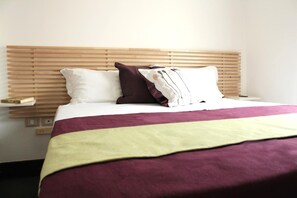 Una habitación de hotel con cabecera de madera, cama con colcha morada y blanca, y almohada blanca.