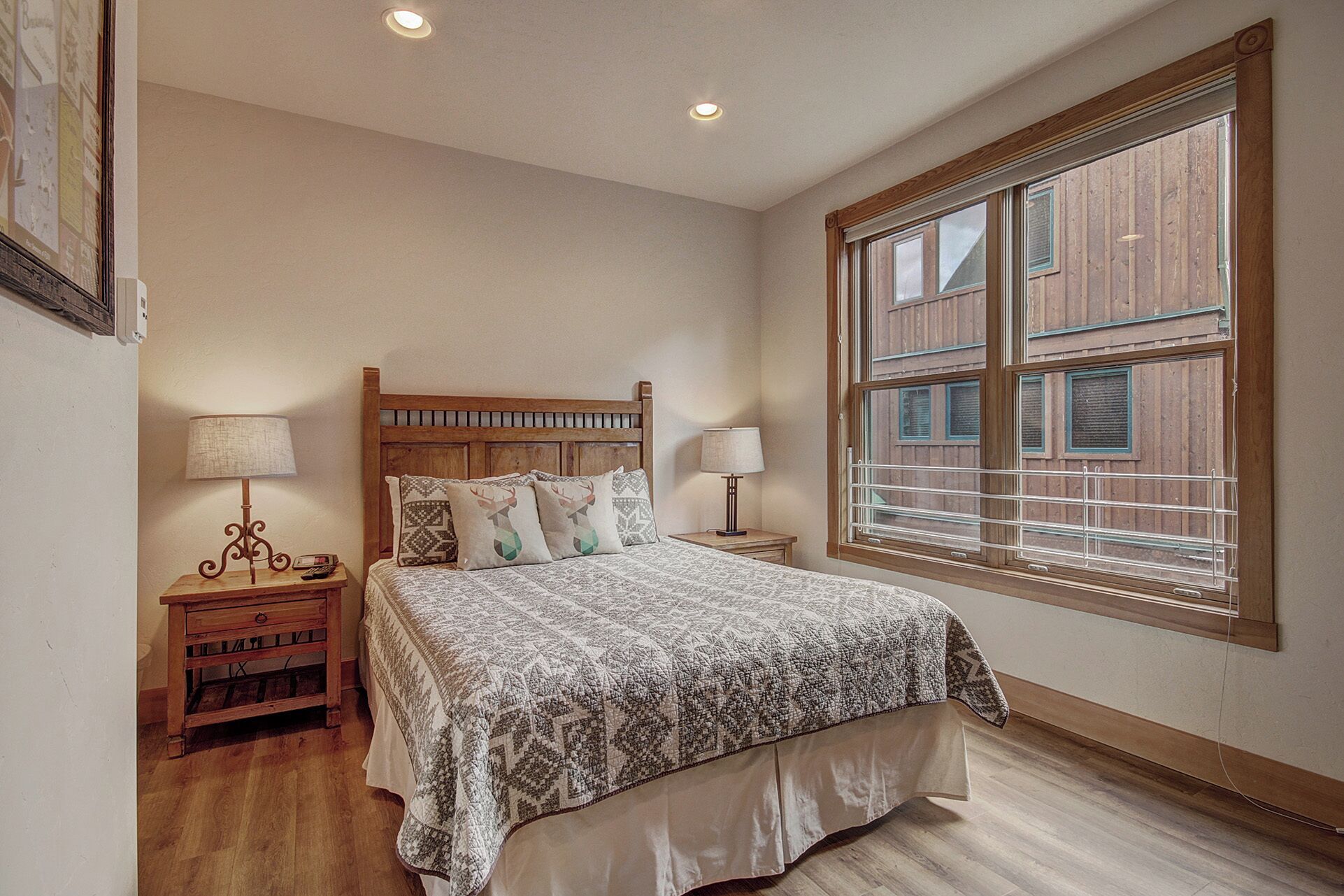 A bedroom with a wooden bed, two bedside tables with lamps, a large window with blinds, and a wooden floor.