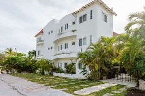 A multi-story white building with balconies, surrounded by greenery and palm trees.
