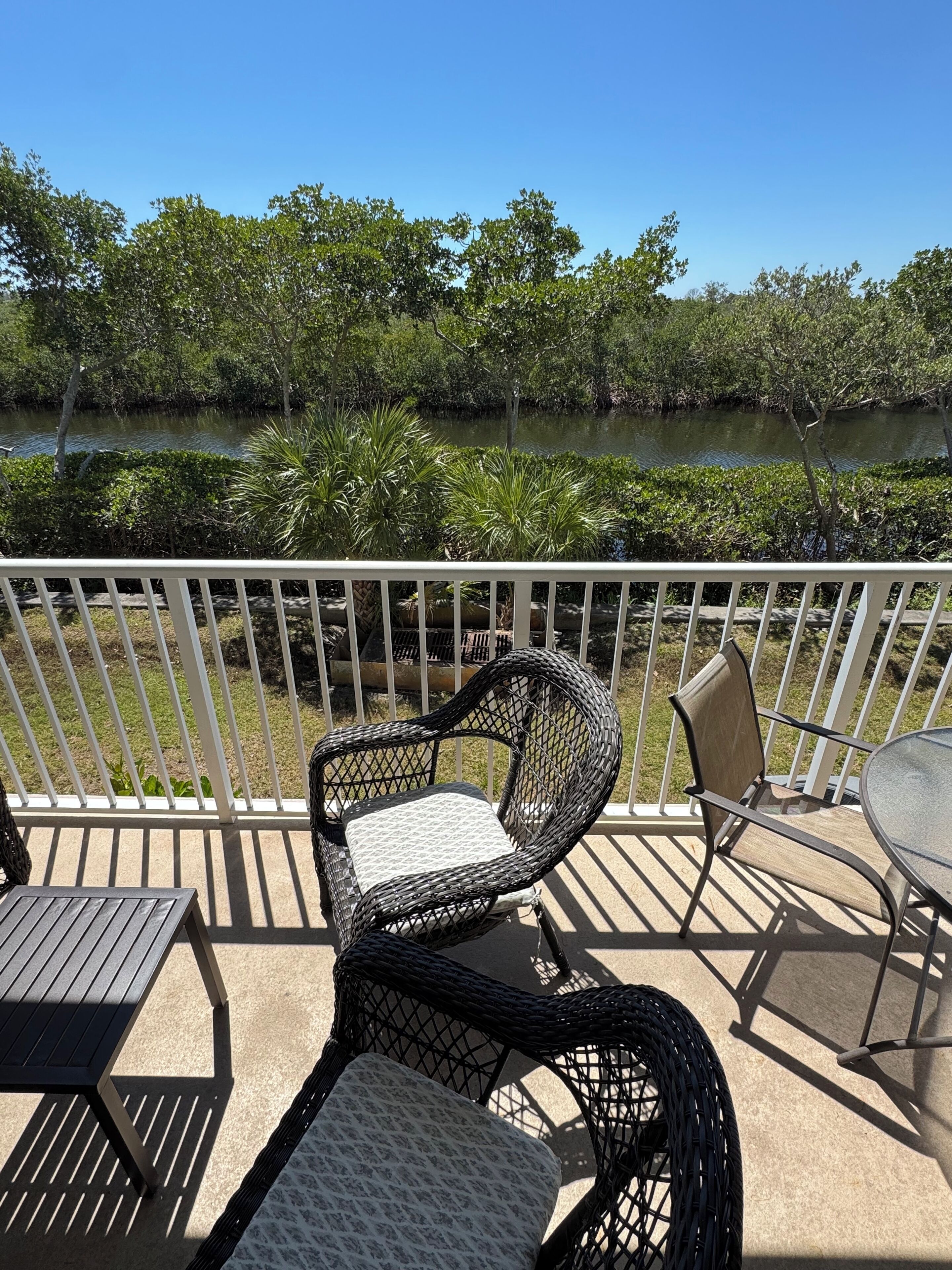A balcony with wicker chairs and a table, overlooking a body of water and greenery.