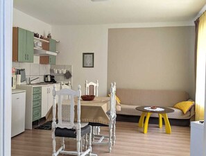 Espaço compacto com cozinha americana, área de jantar com mesa e cadeiras, sofá e uma pequena mesa redonda com um recipiente.