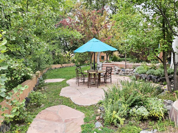 A patio area with a table and chairs under a blue umbrella, surrounded by a garden with various plants and a stone pathway.