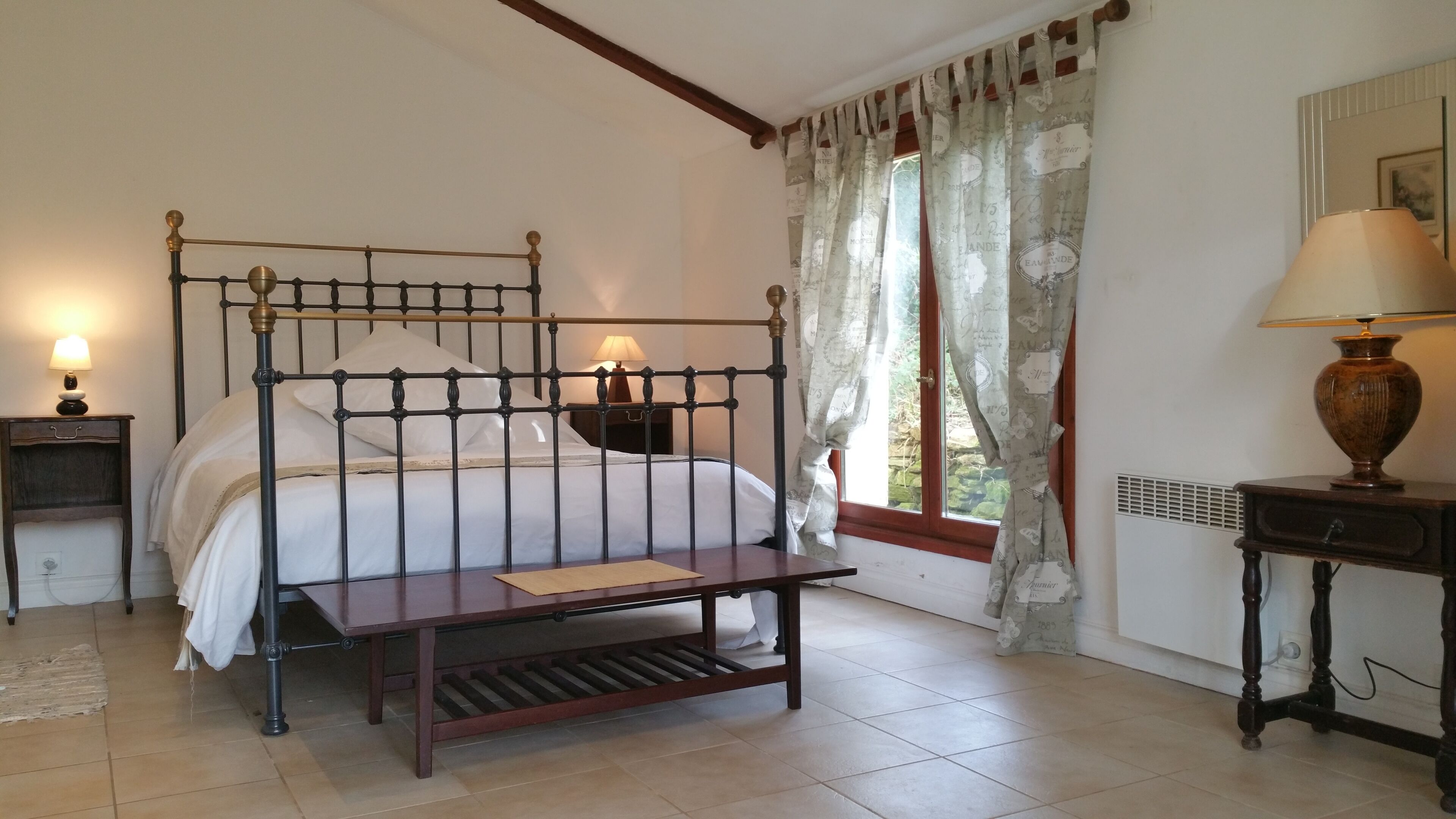 Un dormitorio con cama con dosel, mesitas de noche con lámparas, un banco de madera y una ventana con cortinas.