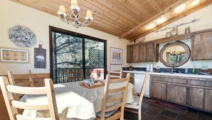 A dining area with a wooden table and chairs, a chandelier, and a kitchen with wooden cabinets and a large mirror. - BEAUTIFUL VIEWS, Cute 1,200 sq ft, HOT TUB! Close to slopes & Lake! FENCED YD (Big Bear Lake)