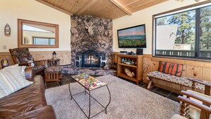 A cozy living room with a stone fireplace, wooden furniture, and a large window offering a scenic view. - BEAUTIFUL VIEWS, Cute 1,200 sq ft, HOT TUB! Close to slopes & Lake! FENCED YD (Big Bear Lake)