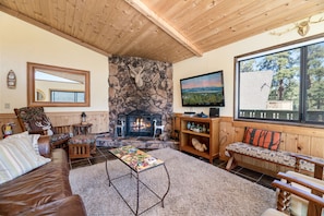 A cozy living room with a stone fireplace, wooden furniture, and a large window offering a scenic view.