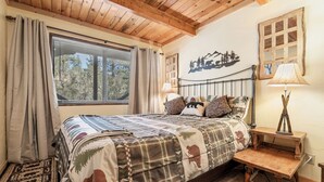 A bedroom with a bed, two bedside tables, a window with curtains, a wall-mounted lamp, and a wall decoration. - BEAUTIFUL VIEWS, Cute 1,200 sq ft, HOT TUB! Close to slopes & Lake! FENCED YD (Big Bear Lake)