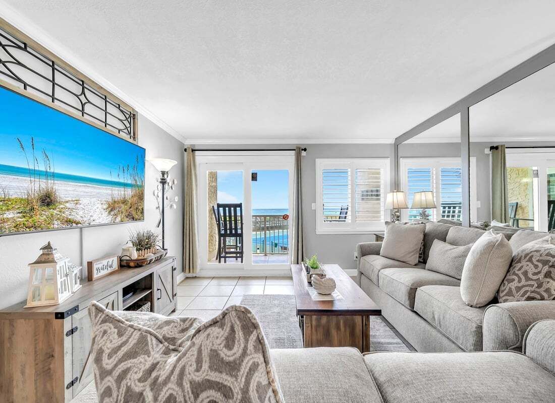 A living room with a sofa, a coffee table, and a painting of a beach scene. There is a balcony with a view of the ocean.