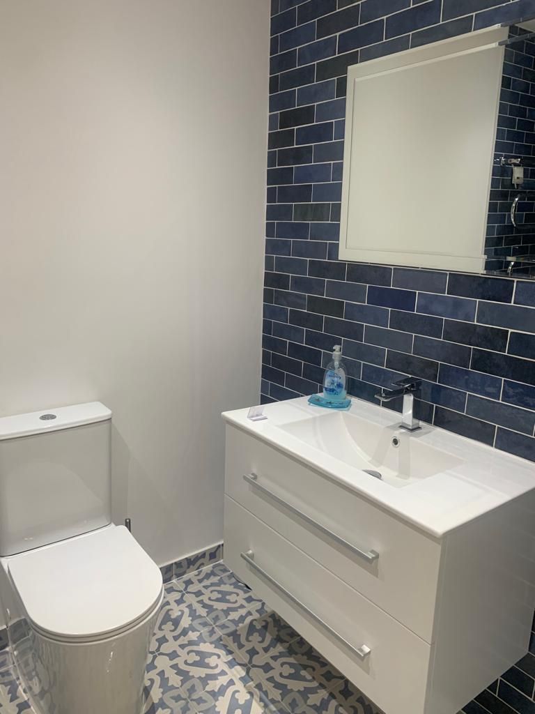 Un baño con inodoro y lavamanos blancos, y un espejo sobre él, además de un piso con diseño.