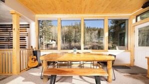 Puinen ruokapöytä ja siihen sopivat penkit huoneessa, jossa on suuret ikkunat, joista avautuu näkymä metsään.
